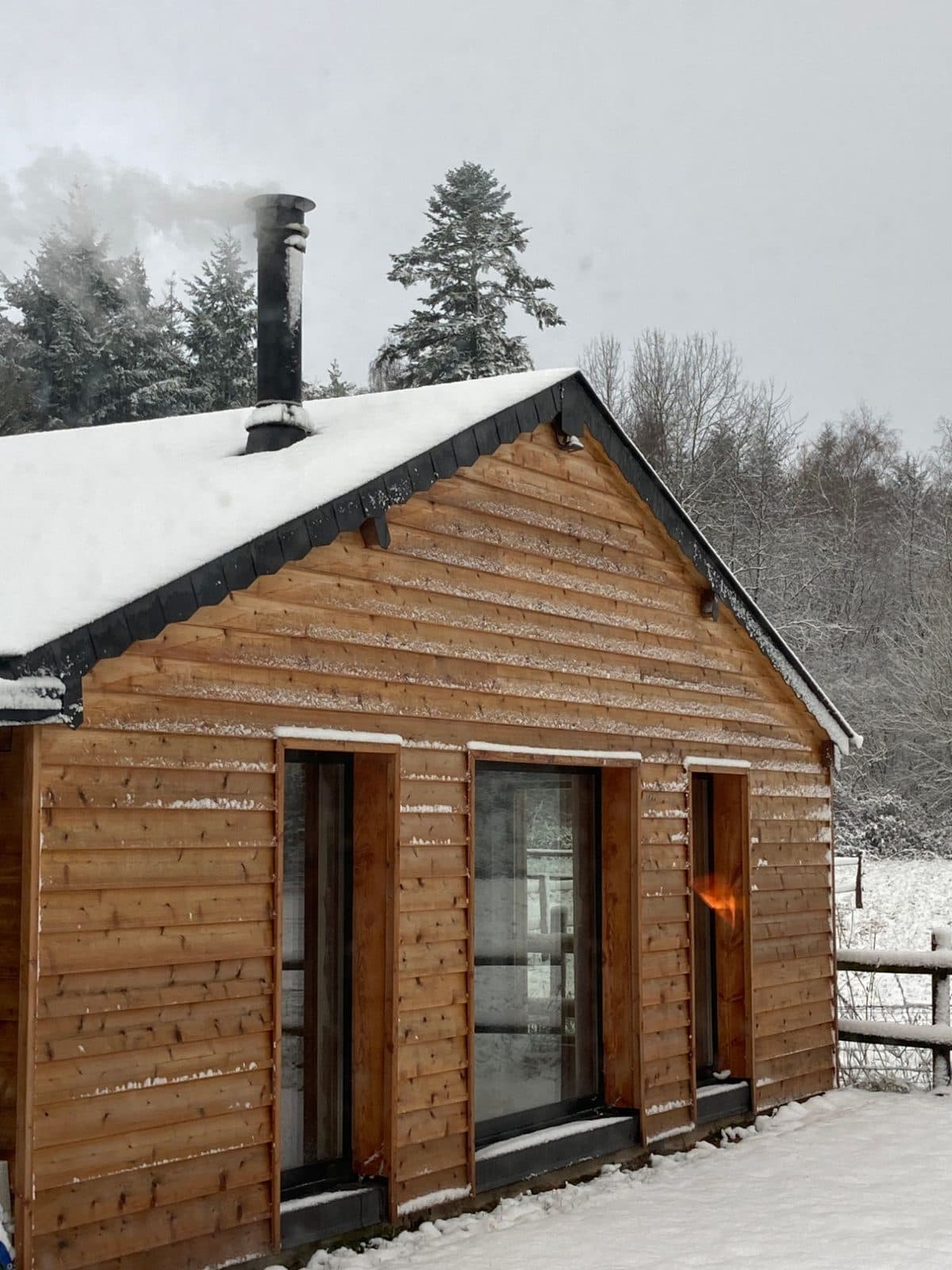 La façade en bois du studio sous la neige, cheminée fumante et forêt enneigée en arrière-plan